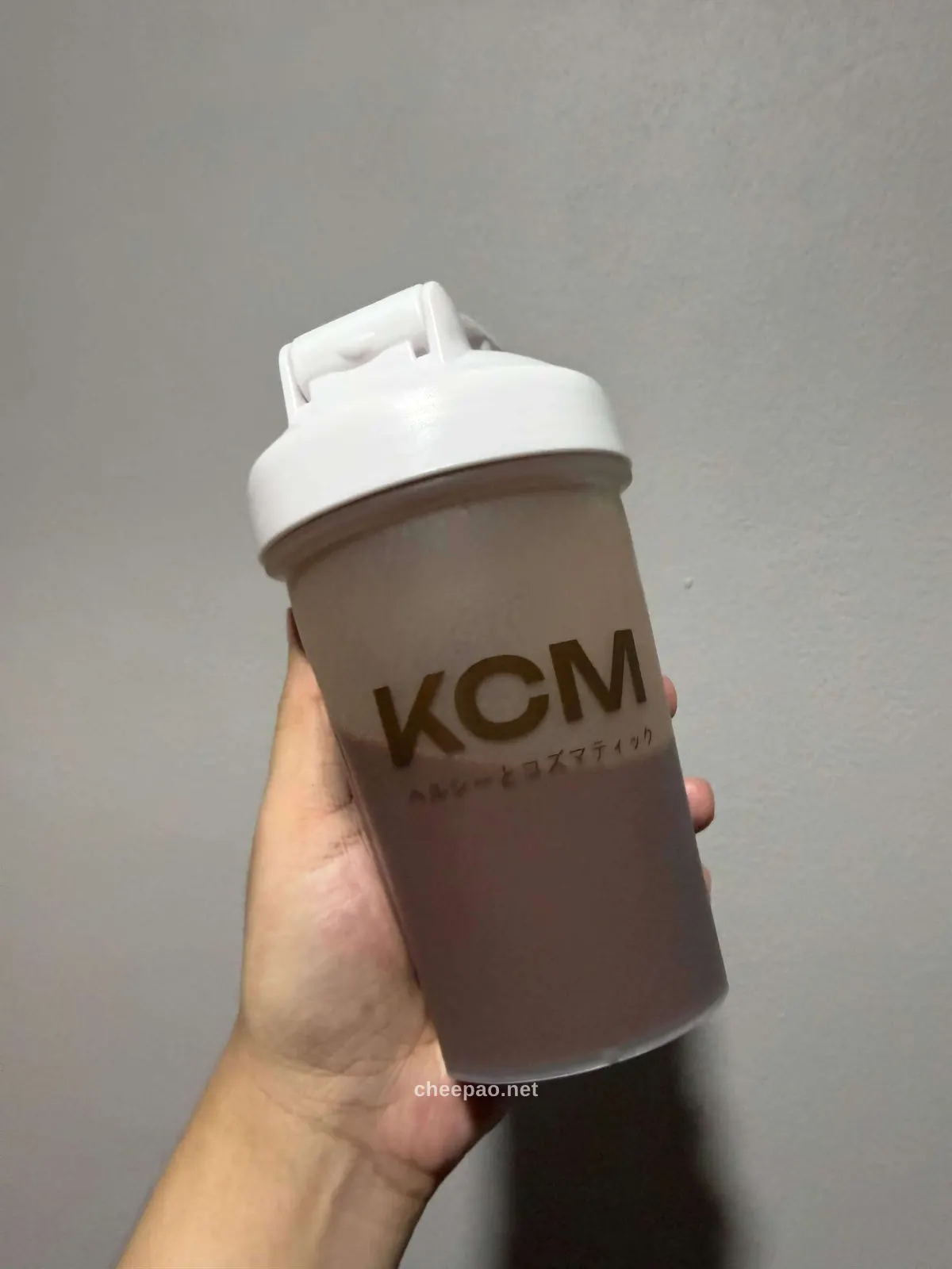 มือถือแก้วเชคเกอร์ KCM ที่ผสมโปรตีนรสช็อกโกแลตแล้ว เห็นเครื่องดื่มสีน้ำตาลนมและโลโก้ KCM บนแก้ว